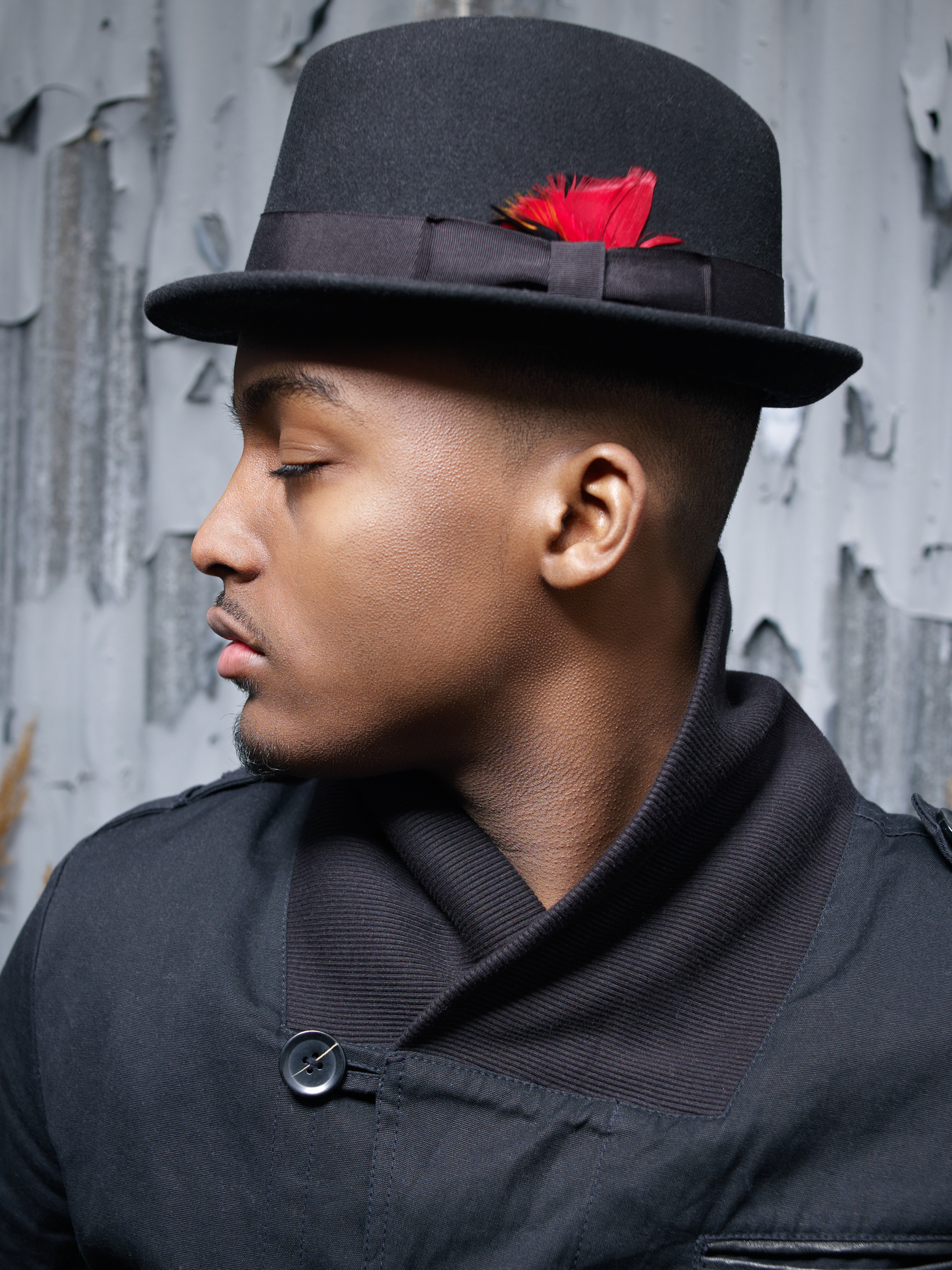 Side profile studio shot of an African American man in a fedora and black jacket with a textured gray background, contemplative mood.
