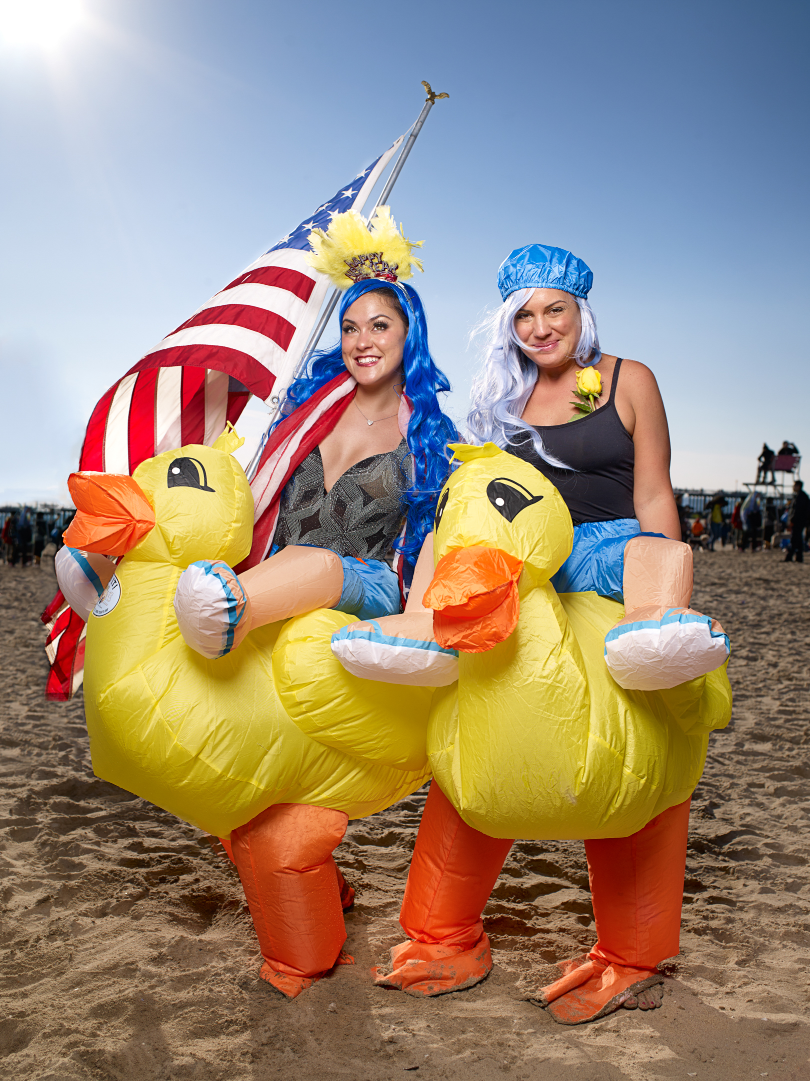 Two women in duck costumes stand on a beach with an American flag in the background, smiling and looking at the camera.