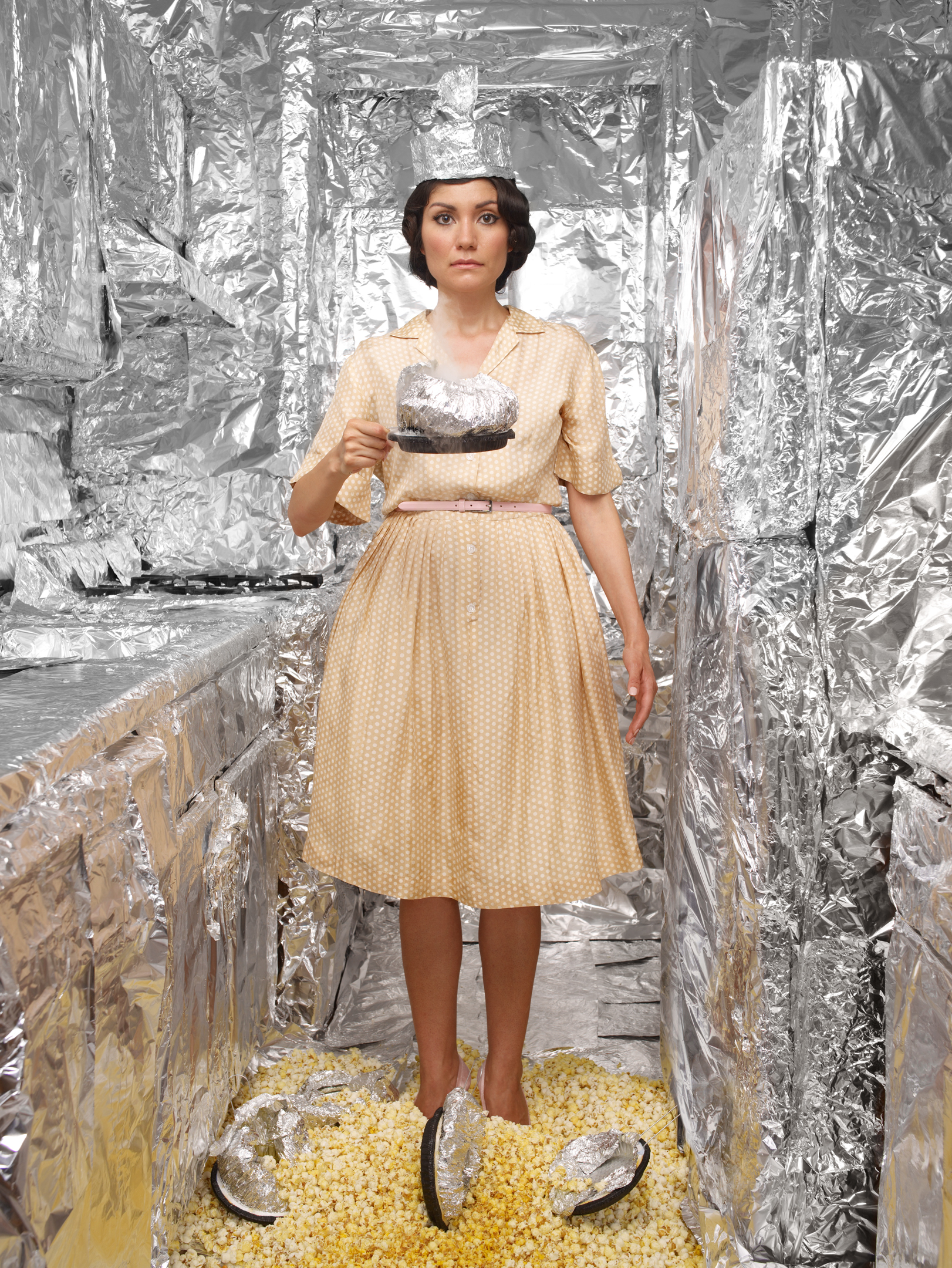 Woman in vintage polka-dot dress holding cake in surreal foil-lined kitchen with popcorn. Conceptual image with retro fashion.
