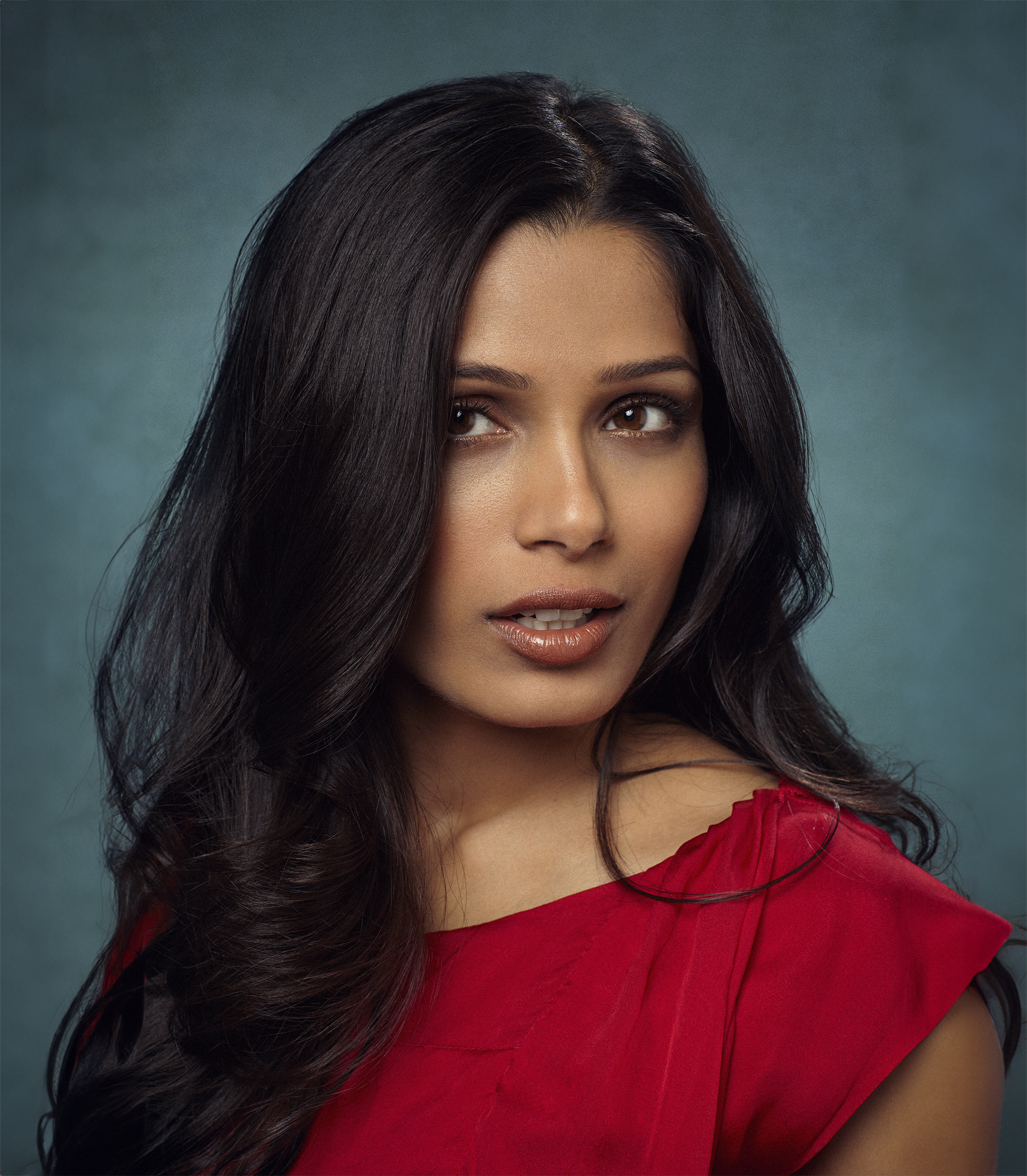 Portrait of a South Asian woman in red dress with long dark wavy hair against a teal backdrop, looking slightly off camera.