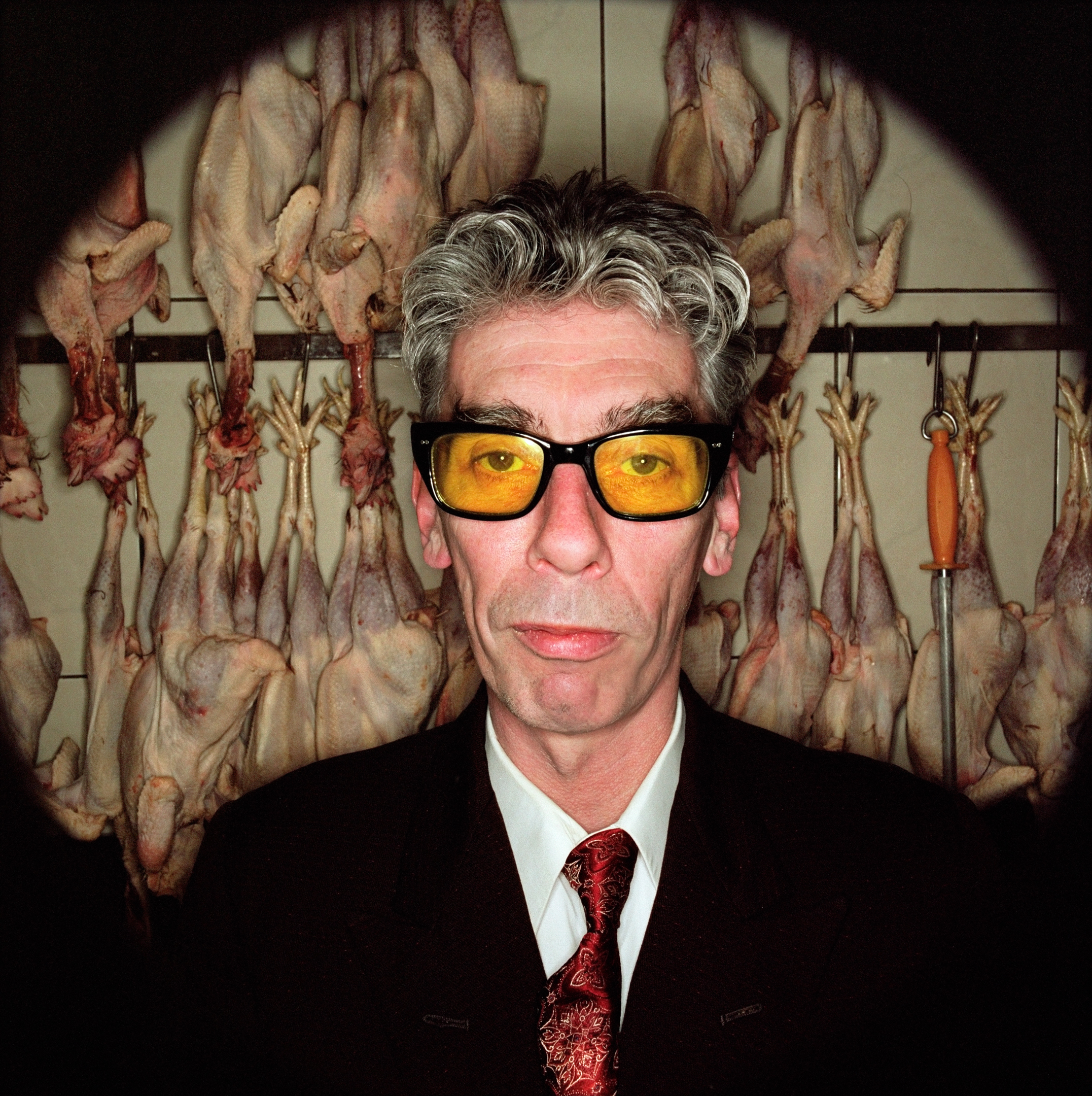 An adult male in a suit and yellow glasses stands in a dark room with hanging raw chickens, creating a surreal and darkly humorous image.