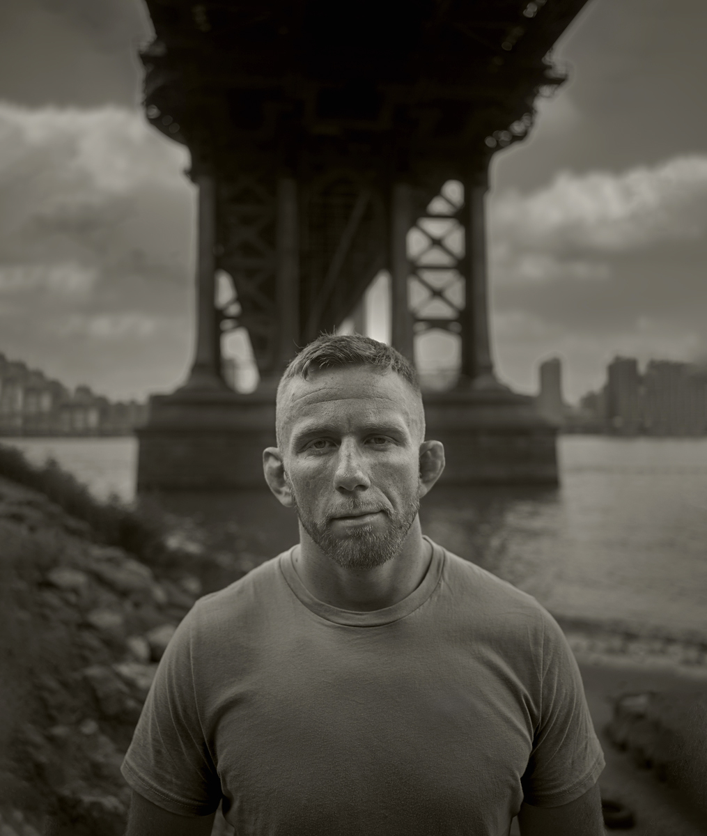 Monochrome portrait of a male adult outdoors, showcasing a gritty aesthetic with a city bridge backdrop and riverside setting.