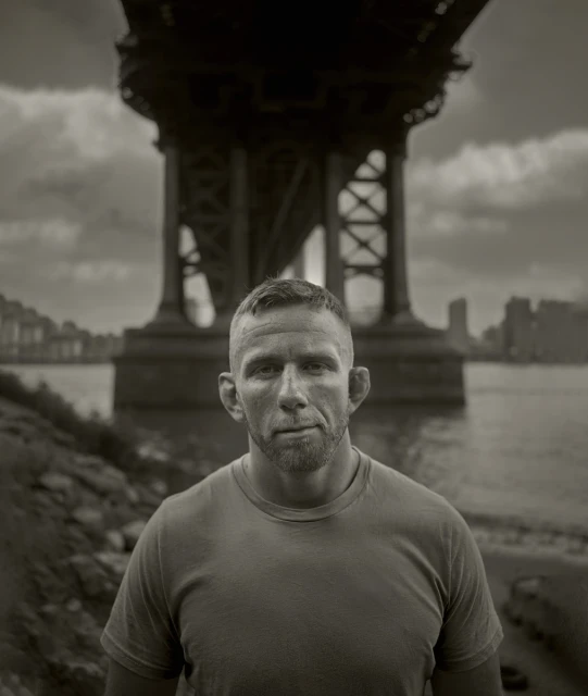 Monochrome portrait of a male adult outdoors, showcasing a gritty aesthetic with a city bridge backdrop and riverside setting.
