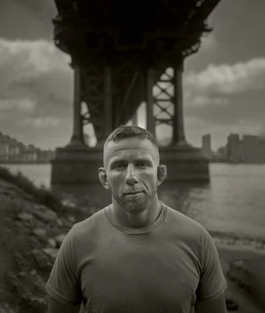 Monochrome portrait of a male adult outdoors, showcasing a gritty aesthetic with a city bridge backdrop and riverside setting.