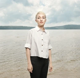 Portrait of a young person with short blonde hair wearing a striped shirt standing by a lake shore.