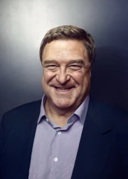 Portrait of John Goodman smiling wearing a blue suit jacket and light purple shirt against a dark textured background