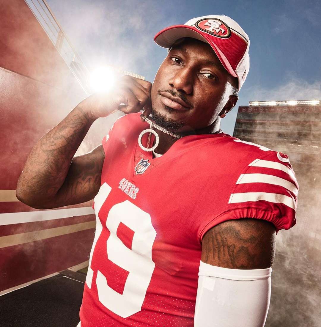 Football player in San Francisco 49ers uniform and hat poses near stadium; red, white, and black athletic fashion and chain jewelry.
