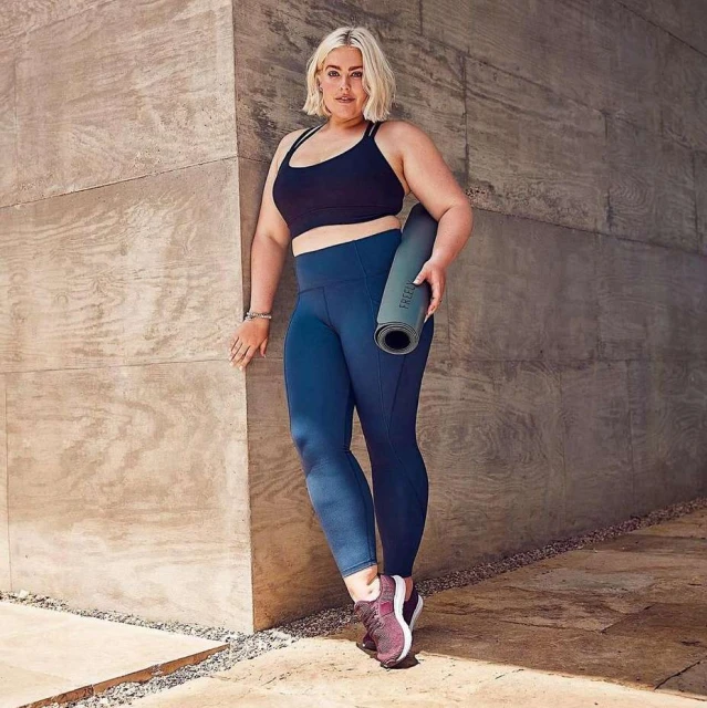 Blonde woman in fitness wear poses against a concrete wall, holding a yoga mat. She wears a black top and blue leggings.