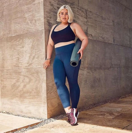 Blonde woman in fitness wear poses against a concrete wall, holding a yoga mat. She wears a black top and blue leggings.