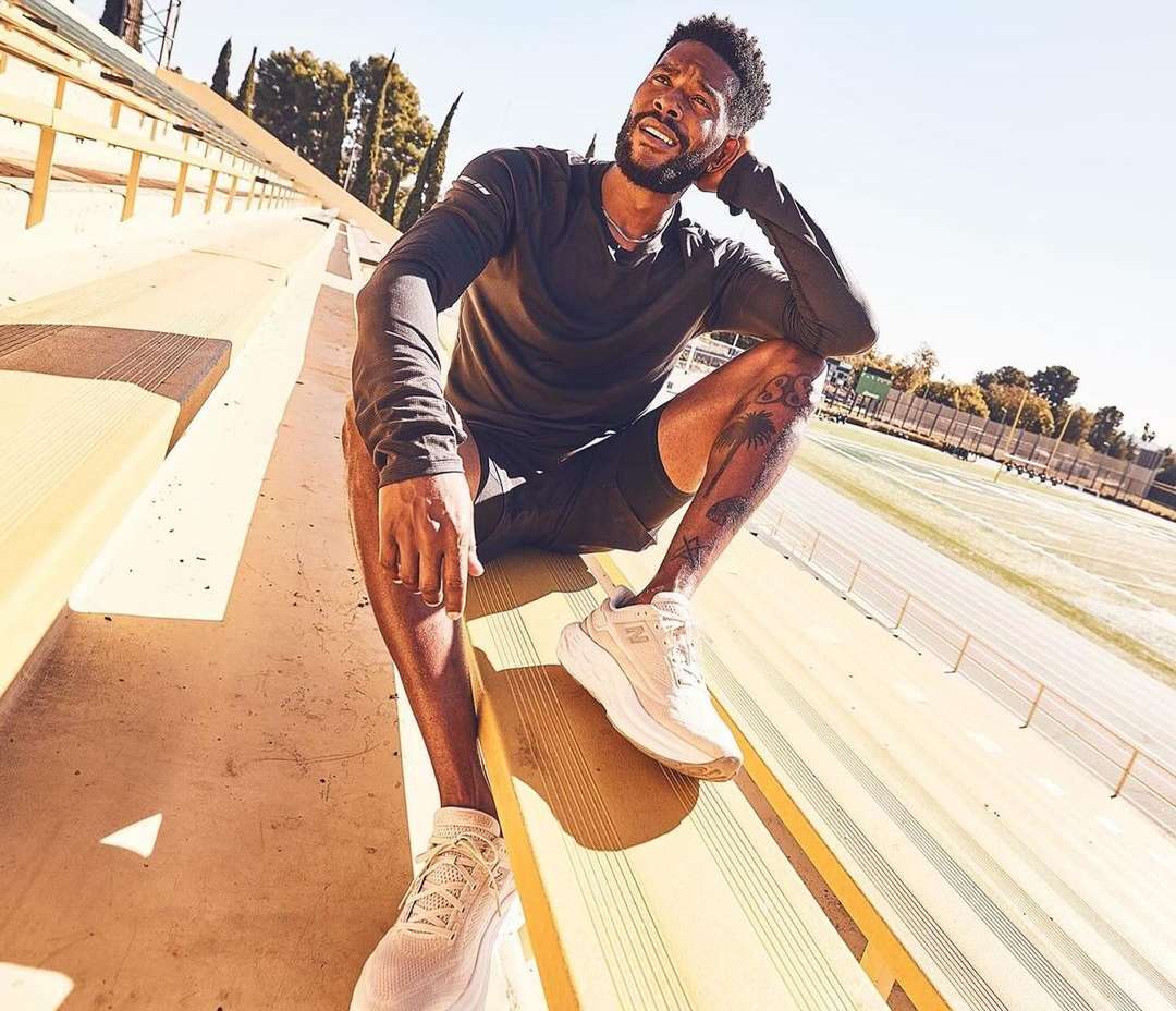 An African-American man sits on stadium seating, wearing a gray long-sleeved shirt, dark shorts, and running shoes in outdoor lighting.