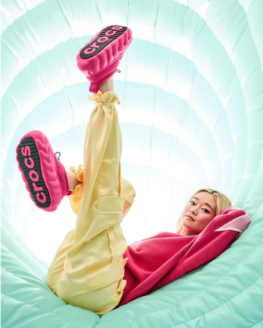 A woman in pink sweater and yellow pants poses in a light blue tunnel with pink Crocs shoes pointed up, showcasing the soles.