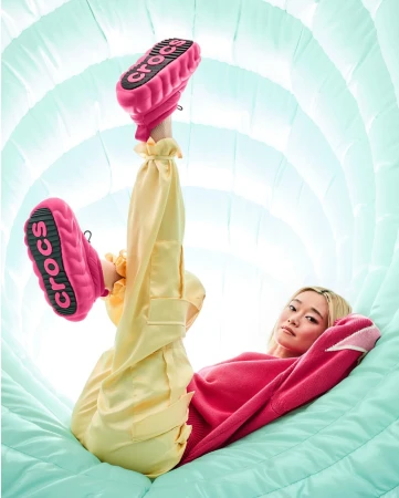 A woman in pink sweater and yellow pants poses in a light blue tunnel with pink Crocs shoes pointed up, showcasing the soles.