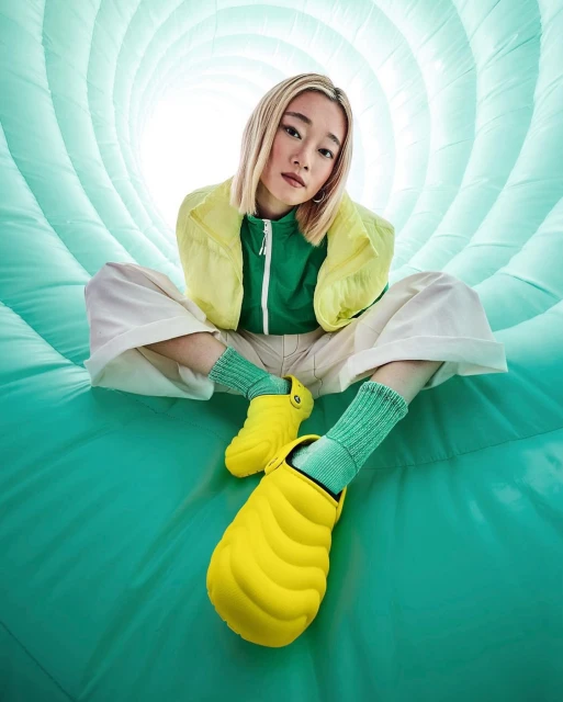 Blonde woman sits in a vibrant aqua tunnel, modeling bright yellow Crocs with green socks, a green top, and a yellow windbreaker.
