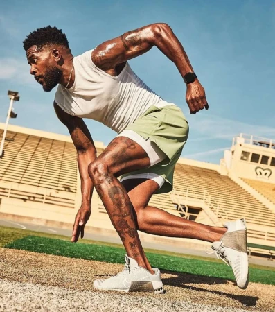 Athletic man running outdoors on a track in a stadium, wearing a white tank top and green shorts on a sunny day.