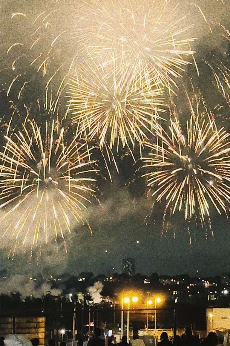 Nighttime fireworks display over a cityscape. Golden and white explosions fill the sky, viewed from a ground-level perspective with silhouetted figures.