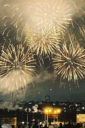 Nighttime fireworks display over a cityscape. Golden and white explosions fill the sky, viewed from a ground-level perspective with silhouetted figures.