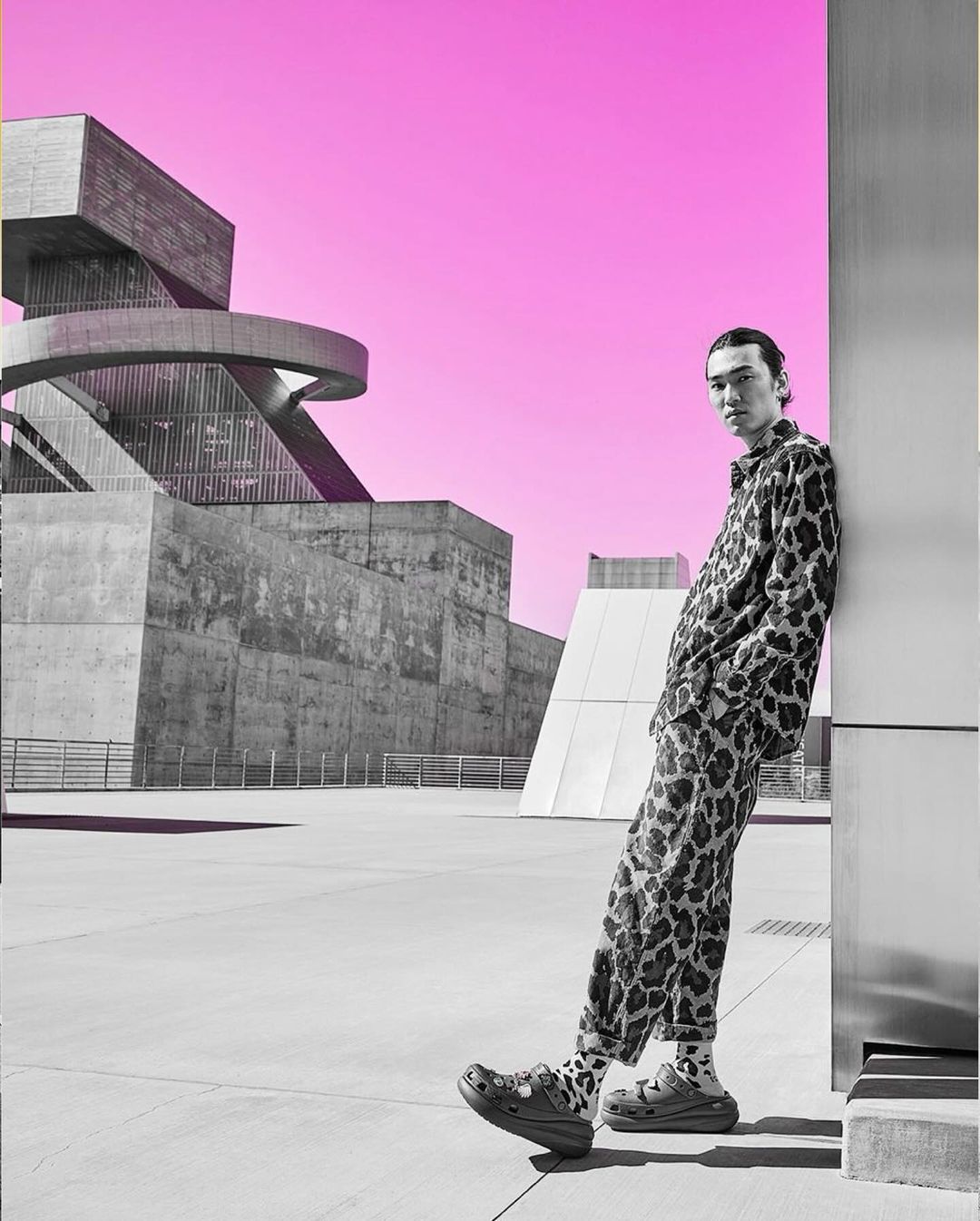 Man poses in animal print suit near modern architecture against a pink sky. Crocs footwear visible. Fashion editorial with urban setting.