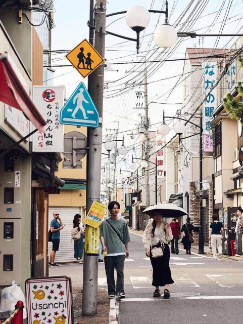 People walk down a Japanese city street on a sunny day, with pedestrian signs, overhead electrical wires, and traditional buildings.