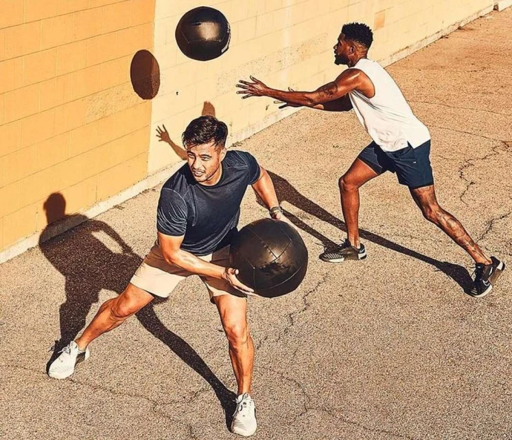 Two athletic men in workout attire are outdoors on a sunny day, throwing a black medicine ball against a yellow concrete wall.