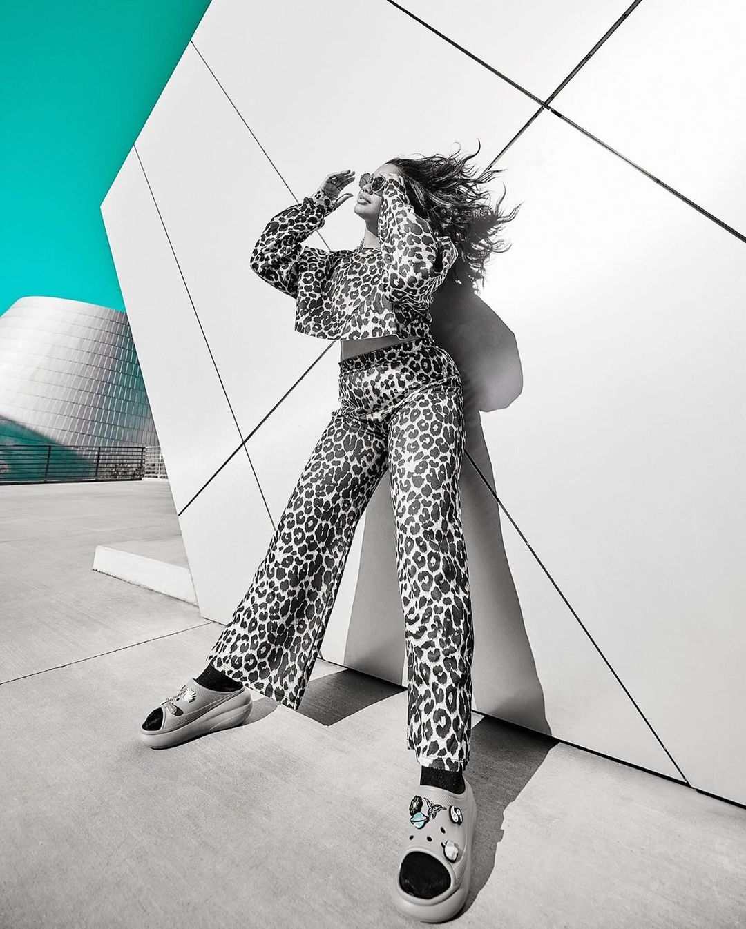 A woman poses outdoors in a printed top and pants, wearing sunglasses, against a concrete wall, with a modern building in the background.
