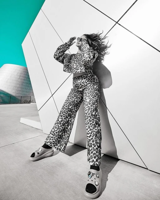 A woman poses outdoors in a printed top and pants, wearing sunglasses, against a concrete wall, with a modern building in the background.