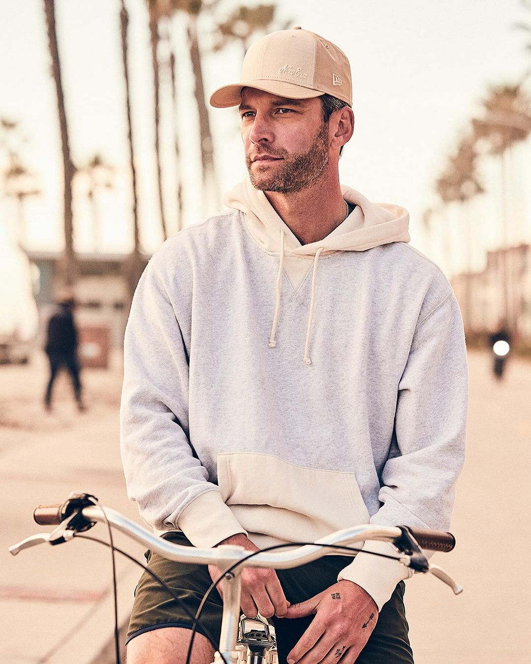 An adult man wearing a hat and sweatshirt poses with a bicycle outdoors near palm trees in soft, diffused lighting on a sunny day.