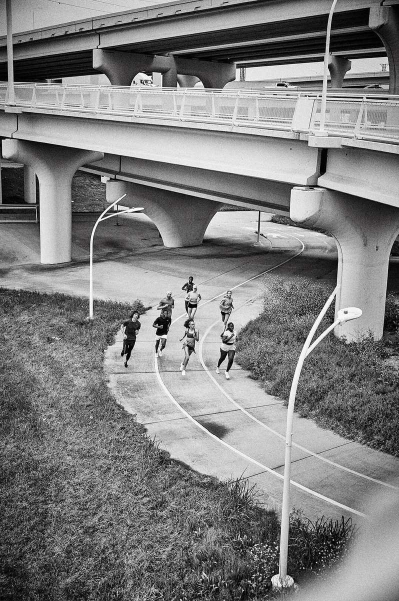 Black and white shot of a group of runners running along a track under an overpass on a cloudy day with vegetation visible.