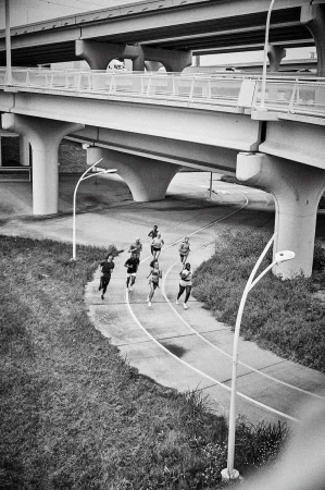 Black and white shot of a group of runners running along a track under an overpass on a cloudy day with vegetation visible.