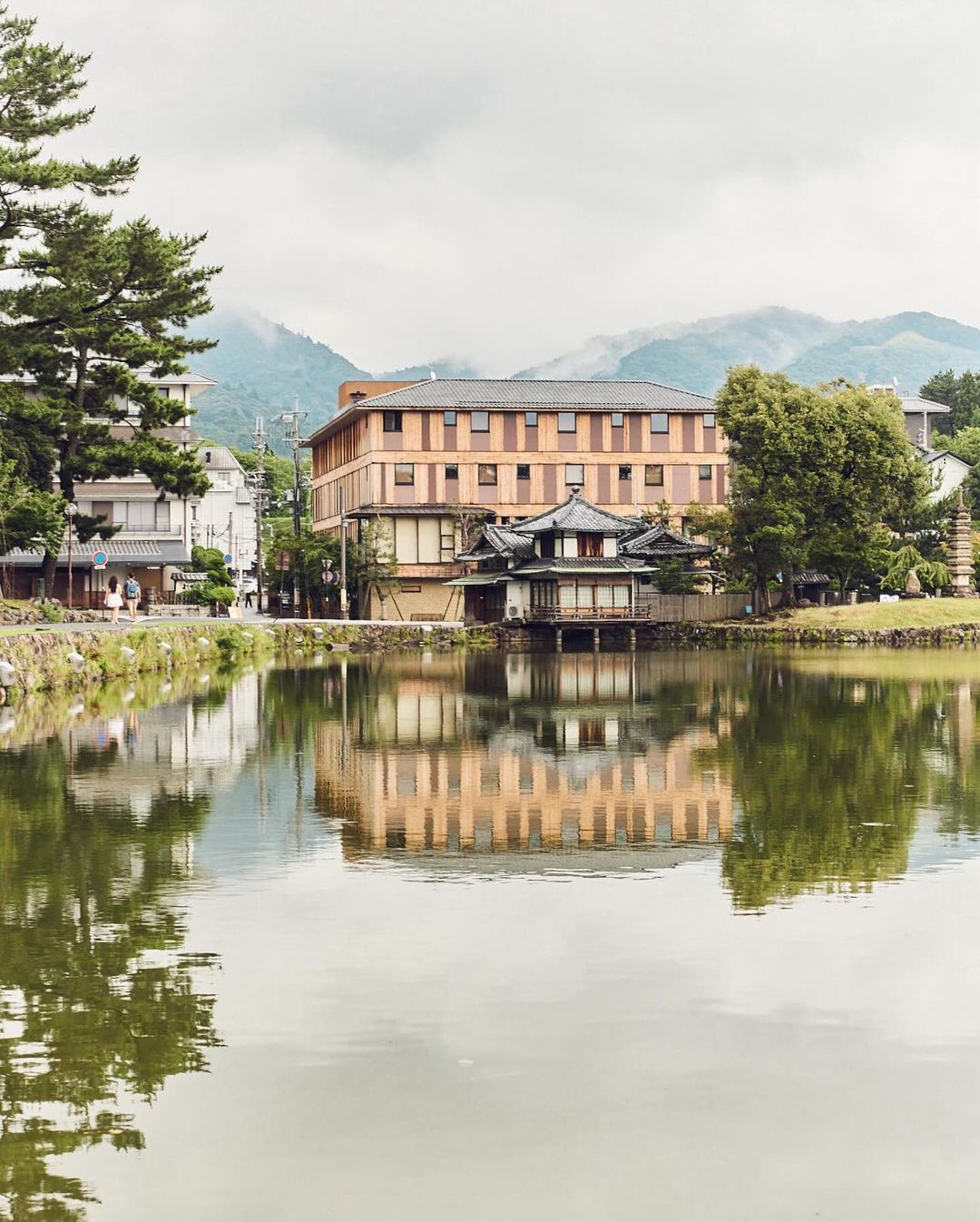 Scenic view of traditional Japanese architecture reflected in a serene lake, with trees and distant mountains under a cloudy sky.