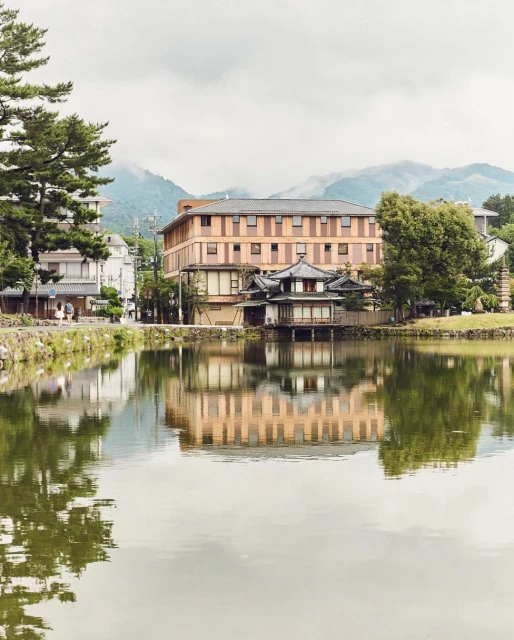 Scenic view of traditional Japanese architecture reflected in a serene lake, with trees and distant mountains under a cloudy sky.