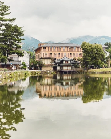 Scenic view of traditional Japanese architecture reflected in a serene lake, with trees and distant mountains under a cloudy sky.