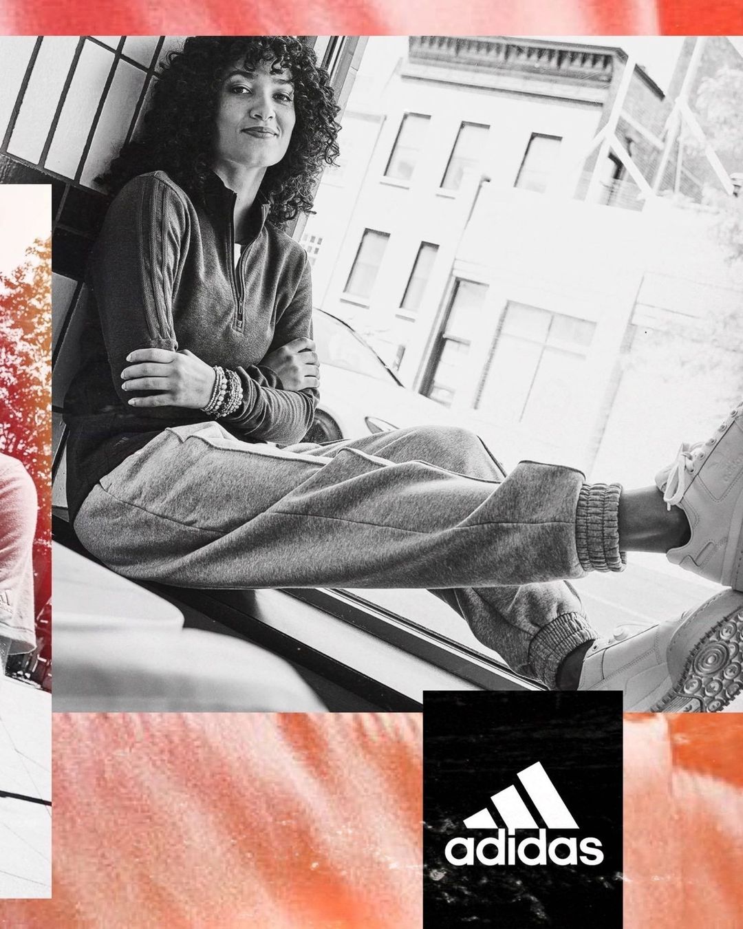 A woman sits in an urban setting wearing grey jogger pants, a quarter zip top, and sneakers, with a building and logo visible.