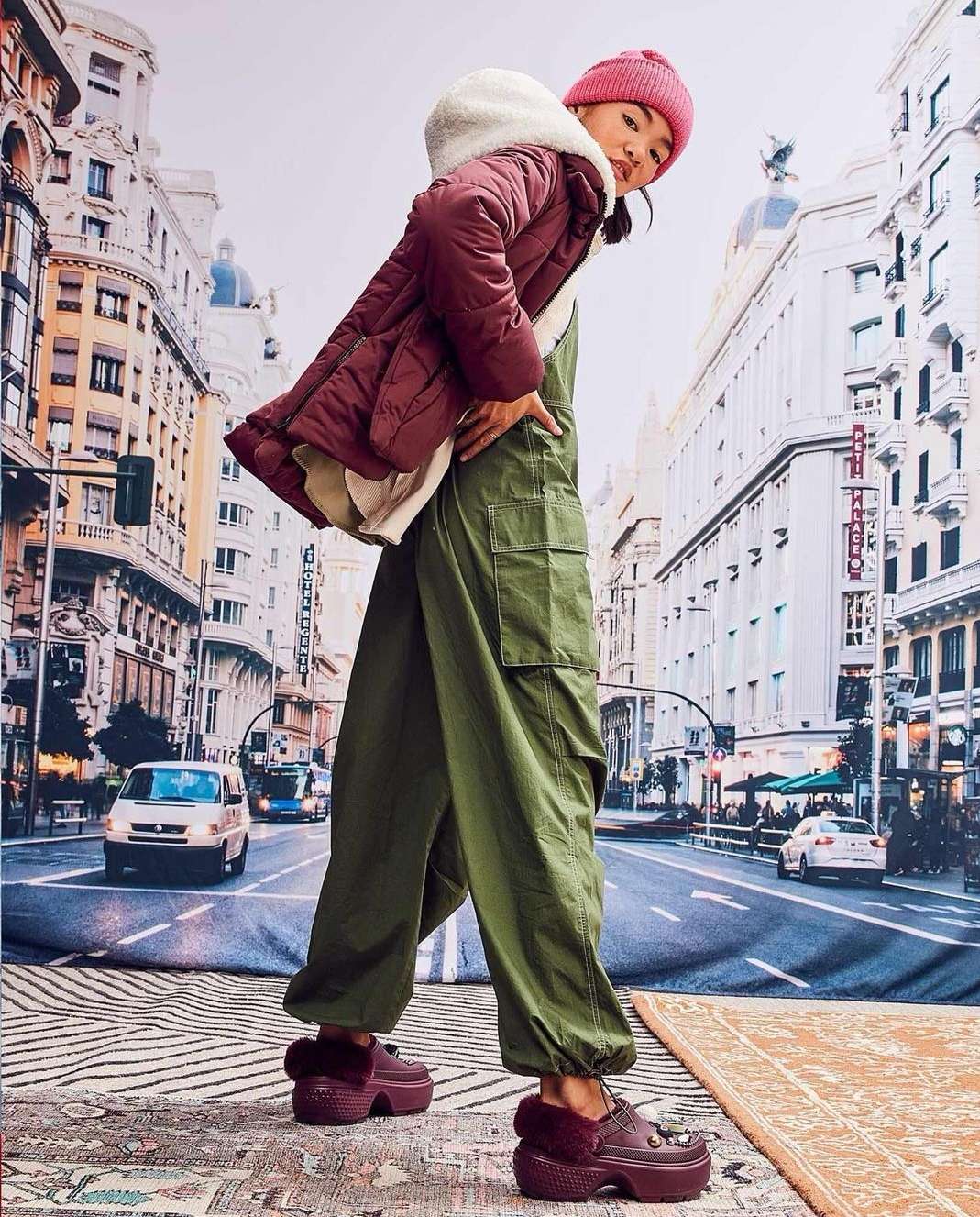 An Asian woman poses in green overalls and maroon platform shoes against a backdrop of city buildings. She wears a maroon coat and hat.
