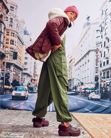 An Asian woman poses in green overalls and maroon platform shoes against a backdrop of city buildings. She wears a maroon coat and hat.