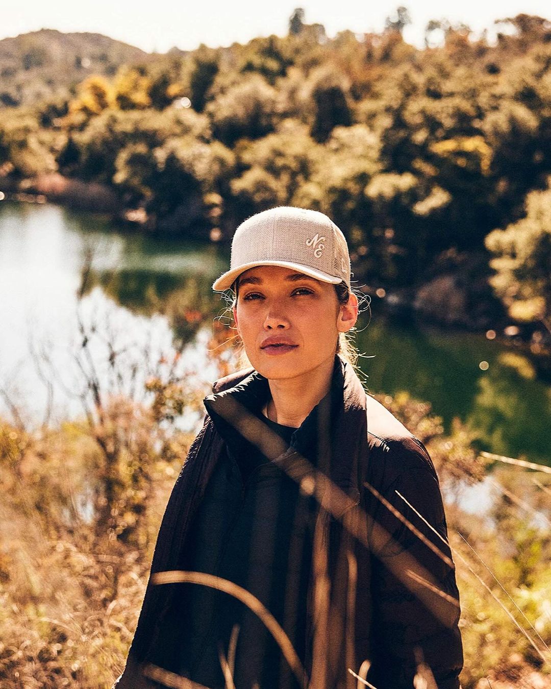 Outdoor portrait of a young adult woman wearing a baseball cap and jacket, standing in a natural setting with a lake in the background.