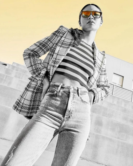 Woman posing in a striped top, jeans, and a plaid blazer, standing on stairs against a yellow sky. Fashion editorial lifestyle shot.