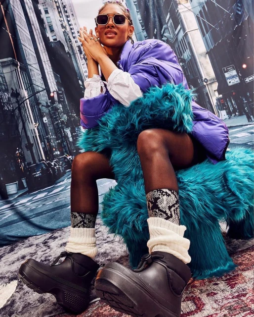Woman in sunglasses wearing a purple puffer jacket, black platform boots, and patterned socks sits against a backdrop of city buildings.