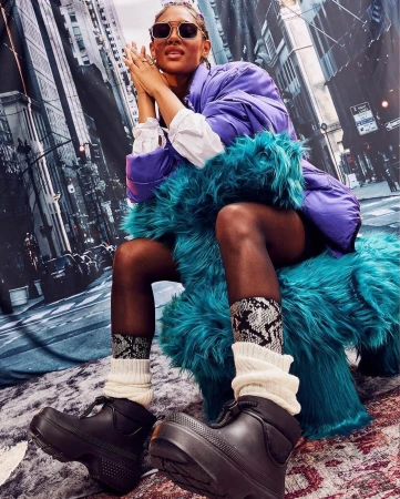 Woman in sunglasses wearing a purple puffer jacket, black platform boots, and patterned socks sits against a backdrop of city buildings.