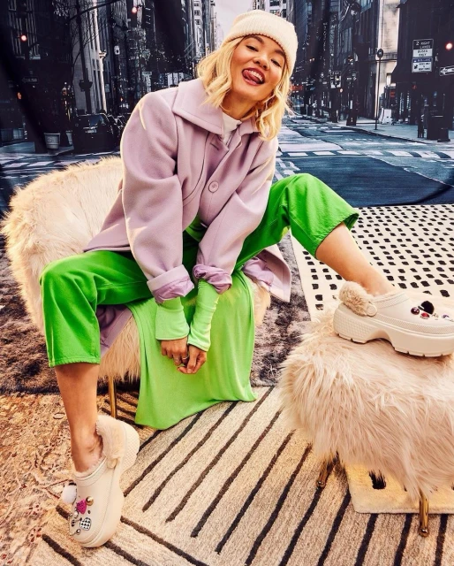 A young woman wearing a lavender coat, green pants, and furry clogs poses in a fur chair in front of a city backdrop.