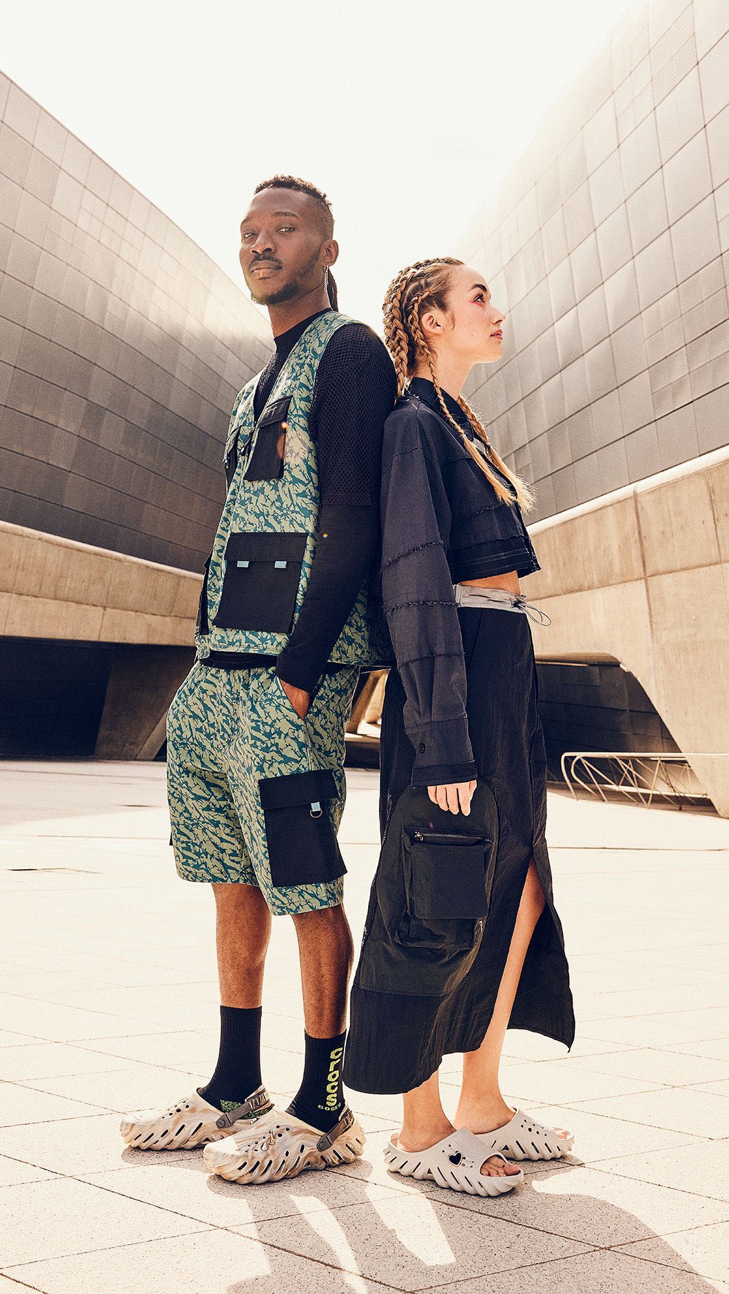 Two fashion models pose outdoors wearing coordinated casual outfits and clogs, standing back to back against an urban backdrop in sunlight.