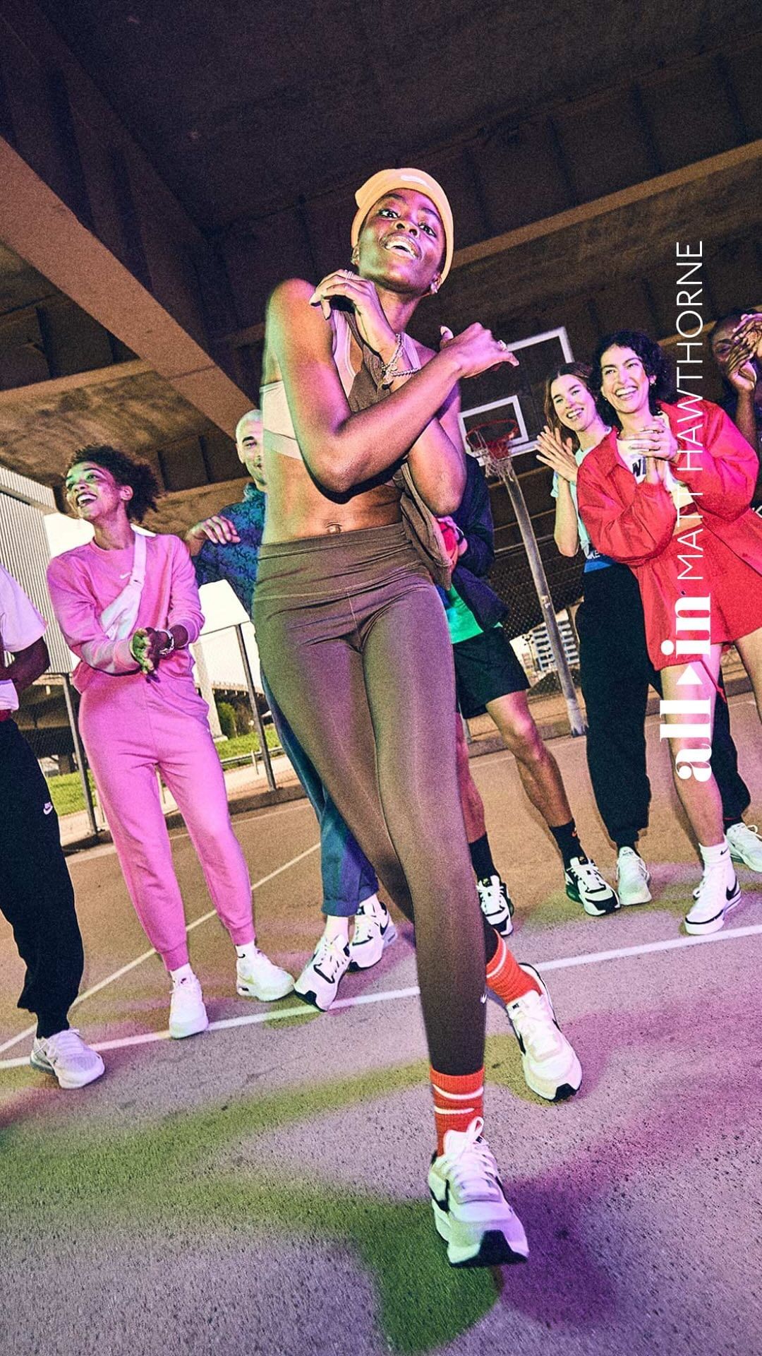 An adult woman in activewear dances with a group of people on a brightly lit outdoor basketball court, wearing athletic shoes and a head covering.