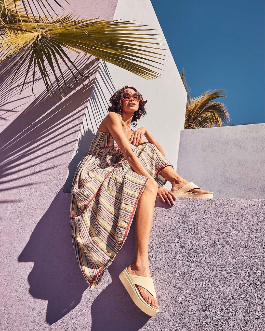 Woman in striped sundress sitting on a stone wall with palm trees against a blue sky. Fashion, travel, and outdoor lifestyle content.