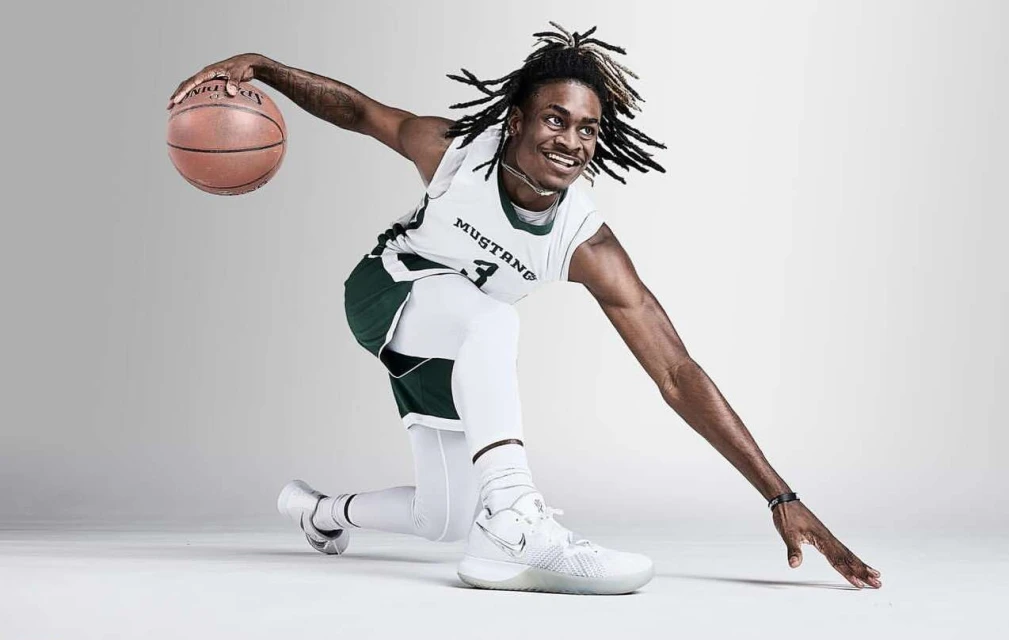 A smiling African American male basketball player with dreadlocks in a white jersey and leggings dribbles a ball on a grey background.