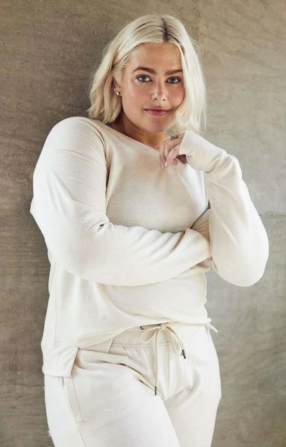A young woman with blonde hair stands indoors, wearing a cream-colored long-sleeve shirt and matching pants, posing against a textured wall.