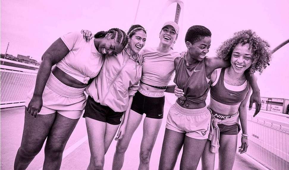 Five smiling women of diverse ethnicities pose together on a bridge in sportswear with the Nike logo visible on their athletic wear.