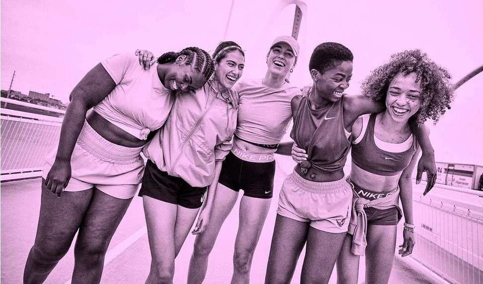 Five smiling women of diverse ethnicities pose together on a bridge in sportswear with the Nike logo visible on their athletic wear.
