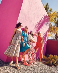 Four women in colorful outfits and sunglasses pose against a vibrant pink architectural wall under bright sunlight.