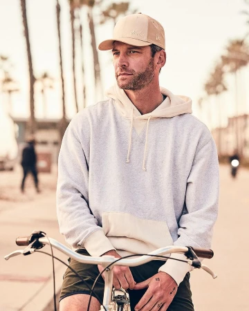 Man wearing a baseball cap and light-coloured hoodie sitting on a bicycle outdoors with palm trees in the background