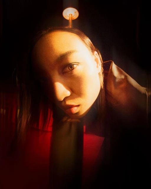 Close-up portrait of a person with warm skin tones, dark hair, and a red jacket, lit by a spotlight in a dark indoor environment.