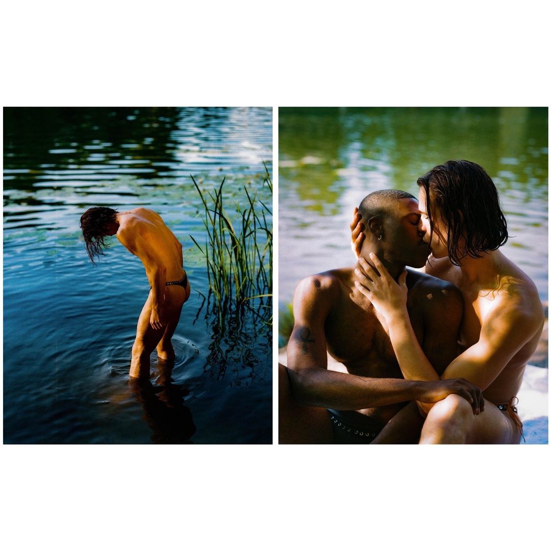 Two men, one standing in blue water with green reeds, the other sitting on the shore, embracing and kissing with lake background.